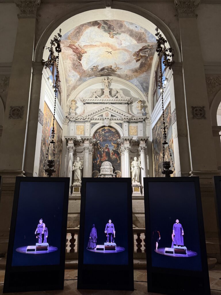 A photo of three vertical black screens showing women standing in a spotlight in a darkened room, arrayed side by side beneath the chapel of a baroque Catholic church with an arched ceiling.