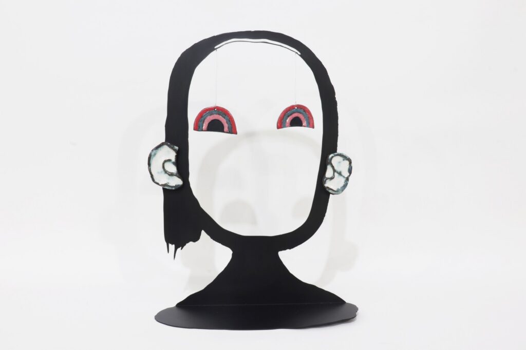 A photo of a cartoon-like sculpture of the outline of a face, with rainbows hanging where the eyes would be.