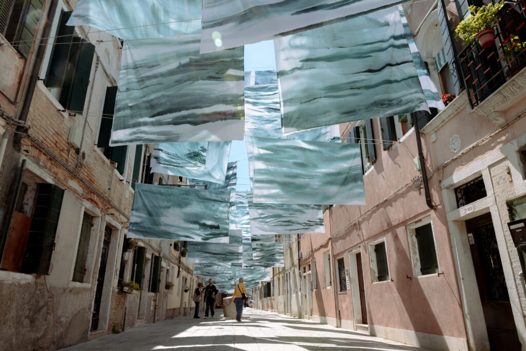 A photograph of translucent blue-green painted sheets suspended between buildings on a sunlit Venetian street as pedestrians walk below.