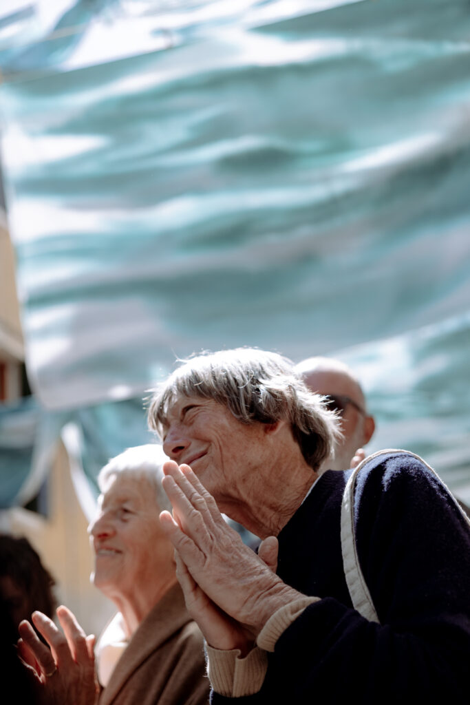 A photograph of two elderly women applauding beneath flowing blue-green fabric installations hanging overhead in Venice.