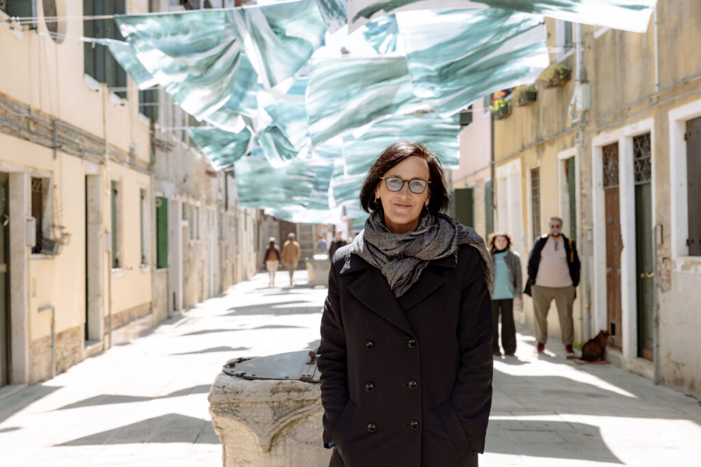 A photograph of a white woman with short dark hair and glasses standing beneath blue-green painted sheets suspended like laundry between buildings on a sunlit Venetian street.