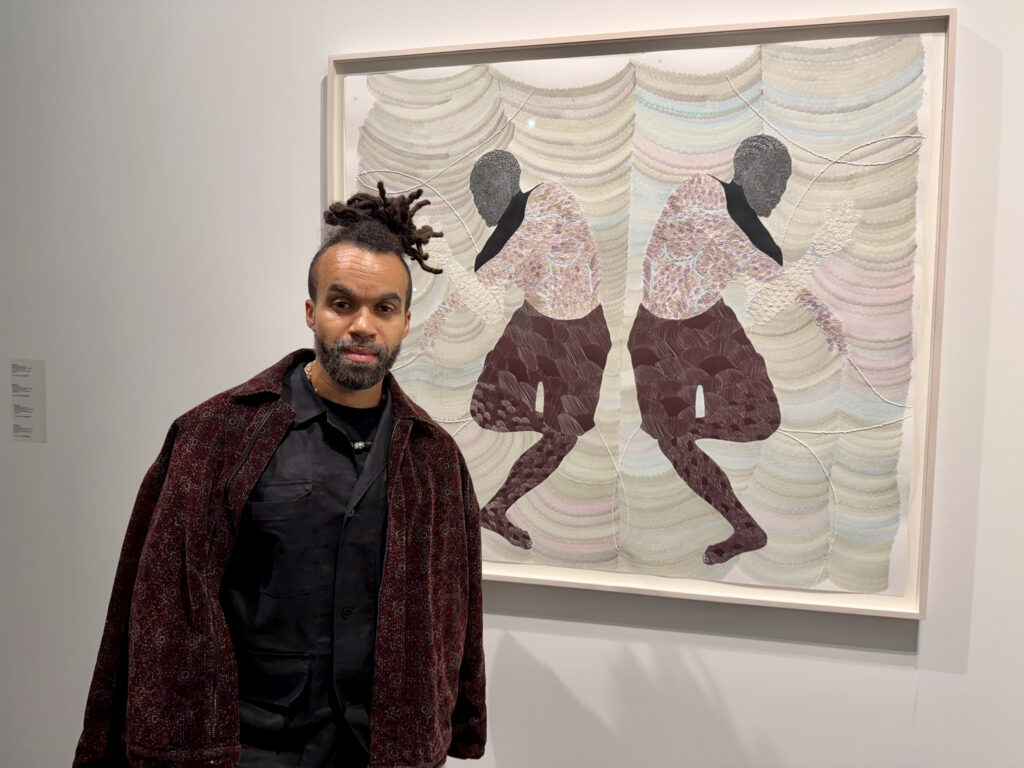 A photo of a young Black man with dreadlocks pulled back into a bun standing in front of a symmetrical, stylized painting featuring two dancers. 