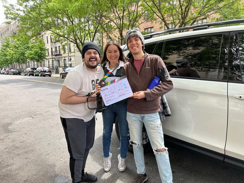 Three young people hold a film clapboard in front of a large white SUV on a city street.