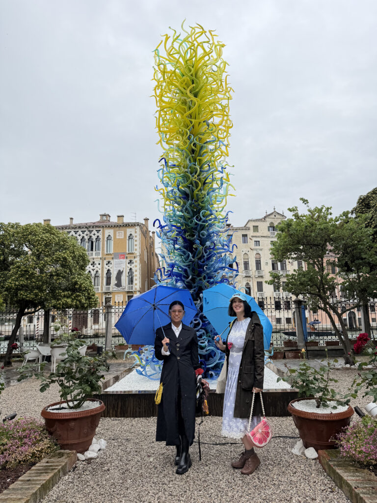 Two women with blue umbrellas stand in front of a tale blue and yellow glass sculpture in a yard in Venice.