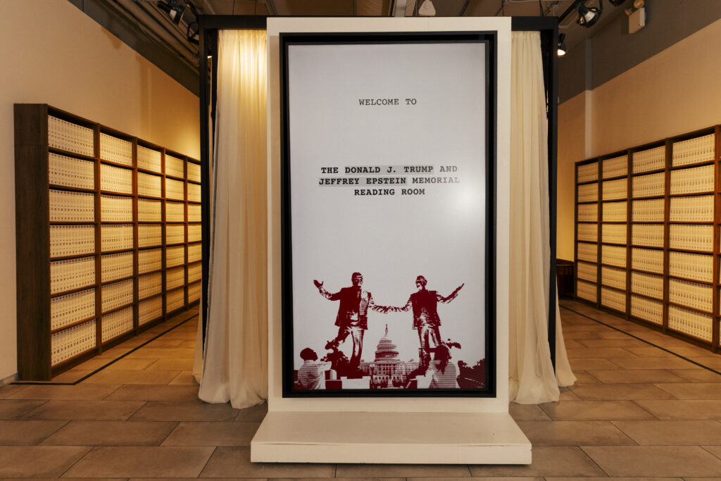A photo of the entrance to the Donald J. Trump and Jeffrey Epstein Memorial Reading Room, with a maroon graphic of the statue of the two men holding hand in front of the U.S. Capitol building framed in the center of the room, and bookshelves filled with printed bound volumes of the Epstein files lining the walls. 