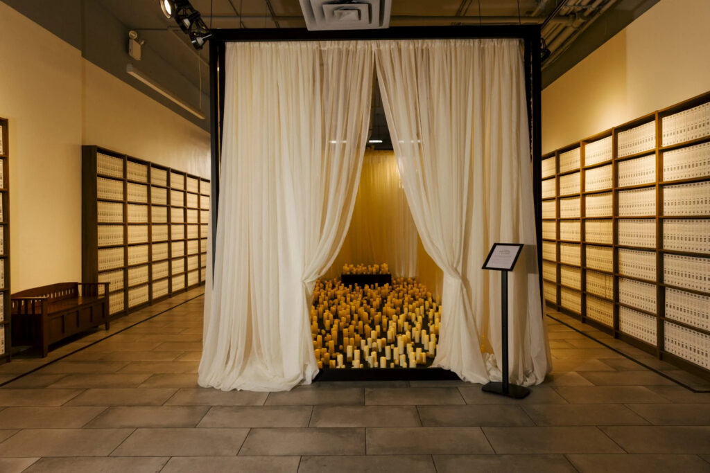 A photo of a galley space lined with bookshelves, and a display of over 1,000 flickering electric candles in the center surrounded by white curtains
