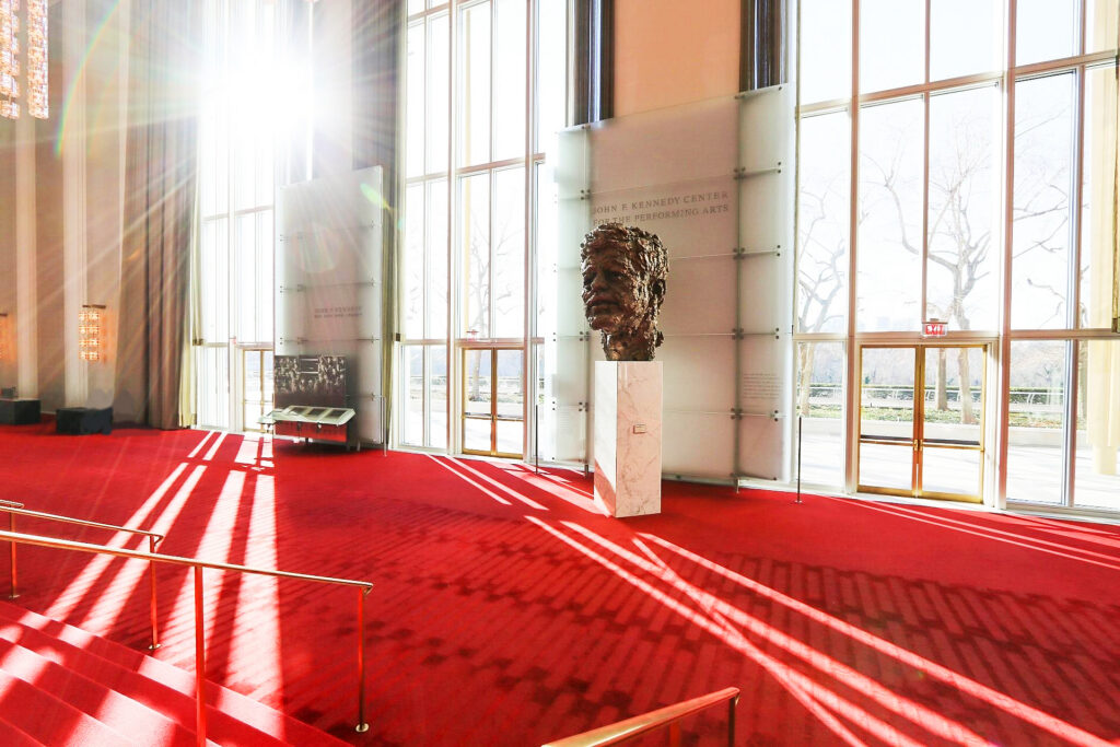 A photo of a large bust of John F. Kennedy displayed on a pedestal on red carpeting, with bright light streaming through the windows behind it.