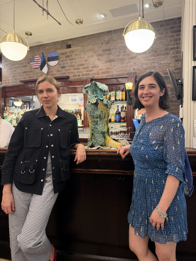 Two women stand at a bar with a large colorful sculpture of a green boot between them. 
