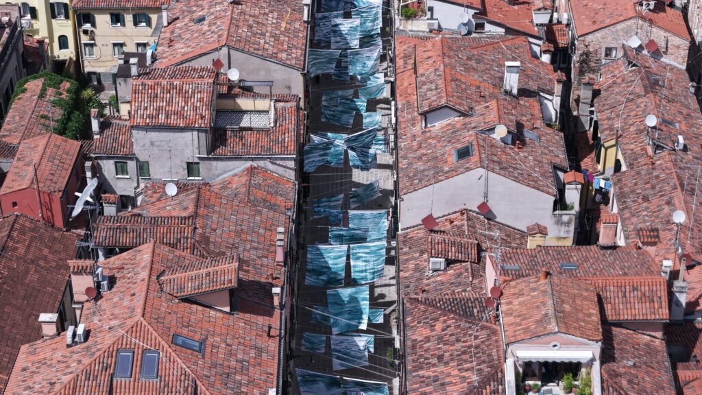 A photograph of painted blue-green bedsheets hanging above a narrow Venetian street lined with terracotta rooftops, viewed from above.