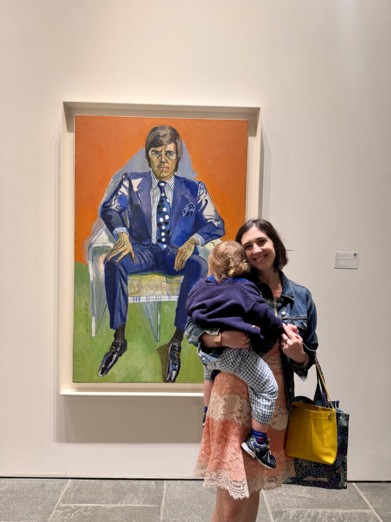 A photo of a woman holding a small toddler who is facing away from the camera. They are standing in front of a colorful Alice Neel portrait of a young man. 