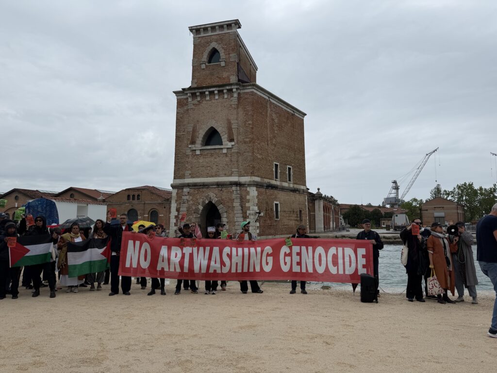 A photo of a group of protesters carrying a large red banner reading "no artwashing genocide" in front of a three story brick tower.