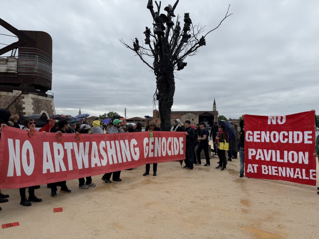 A group of protesters carrying large red banners reading "no artwashing genocide" and "no genocide pavilion at biennale," with a large sculpture of a black tree between the two.