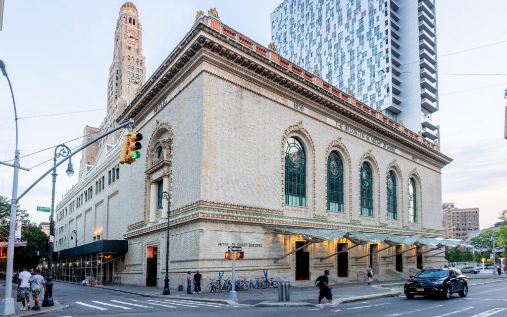 A photo of a Beaux Arts building with a row of tall arched windows on the corner of a Brooklyn street, with a modern skyscraper behind it.