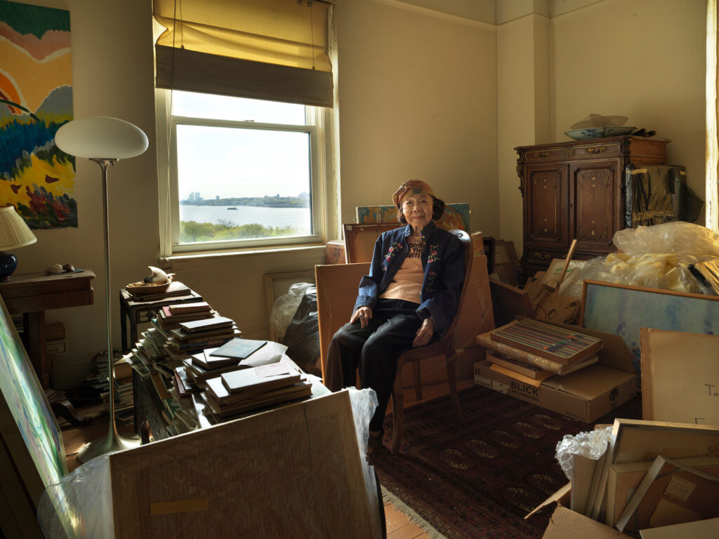 A photo of an elderly woman sitting in her apartment living room with views out on the river, with paintings and books stacked up.