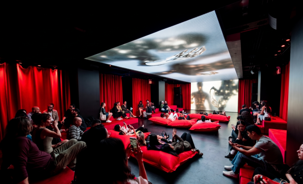 A photo of a room with red curtains on the walls and visitors reclining on red cushions watching a video screen on the ceiling and back wall. 