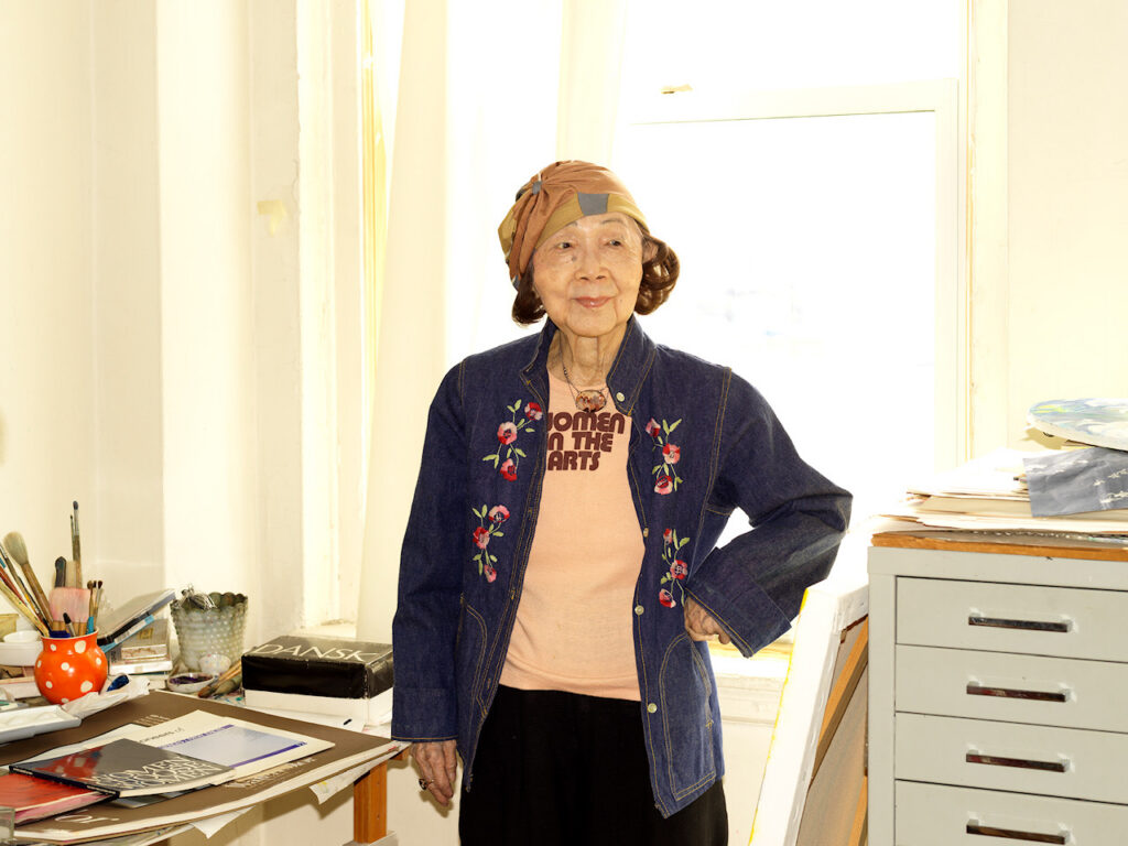A photo of an elderly woman in a cloche hat in a light-filled studio in front of a flat file cabinet and a desk covered in papers and a jar of paintbrushes.