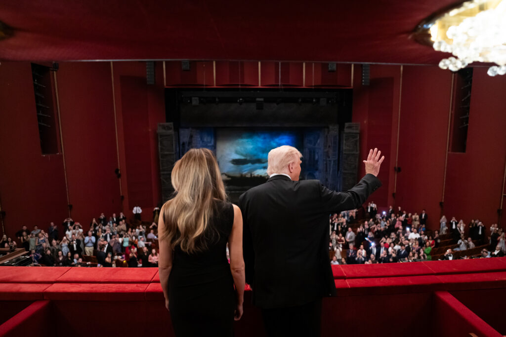 A photo of the backs of President Donald Trump and First Lady Melania Trump waving to the crowds from the balcony of the Kennedy Center Theater. 