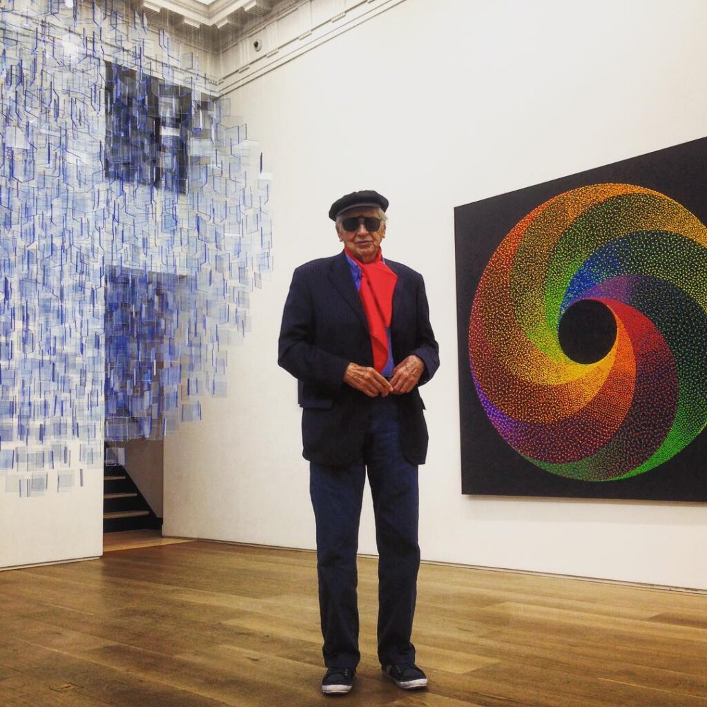 A photo of an elderly male artist in a black suit and hat and sunglasses with a red shirt, standing in an art gallery between a painting of a rainbow gradient in a spiraling circle against a black background and a large hanging mobile of small, translucent blue plexiglass squares hung to make a large circular form. 
