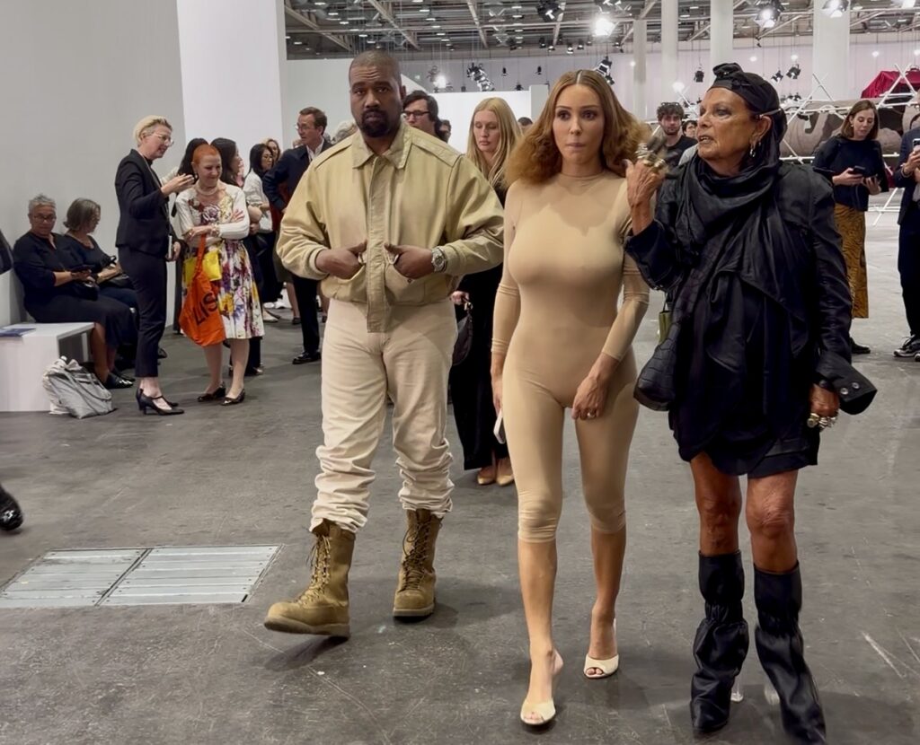 A photo of Kanye West and Bianca Censori in tan outfits, hers a skintight body suit, walking through an art fair with Michèle Lamy, wearing all black.