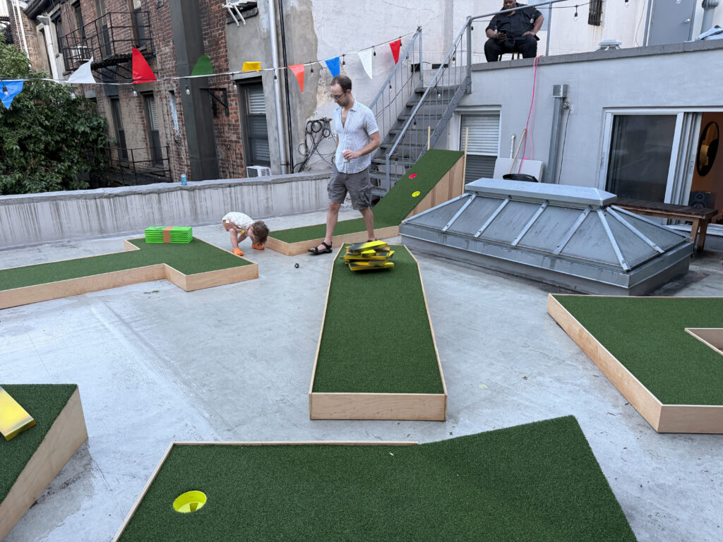A photo of a mini golf course on the roof of an apartment in New York, with a father tending to a toddler who is bending over to play with a ball. 