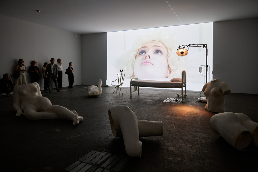 A photo of a film screening in a darkened room, with a close up of a woman's face lying down on a bed in a brightly lit room. In the screening room, a bed and white plaster body part sculptures are in front of the projection, with a small audience standing to watch.