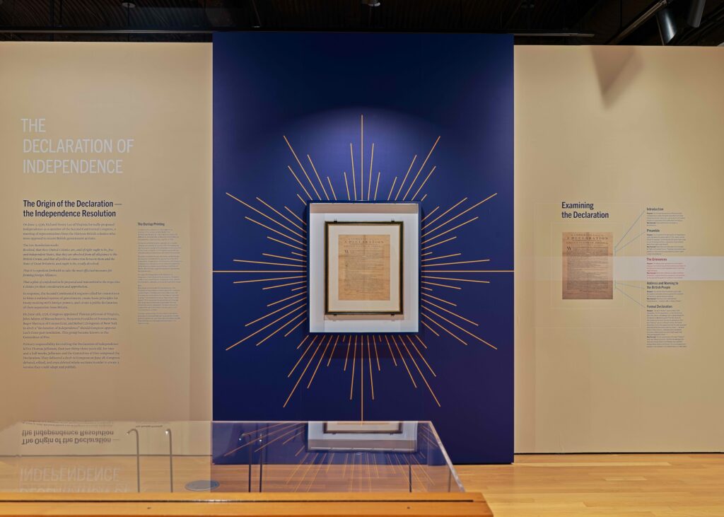 A photo of a museum display with a framed copy of the Dunlap Broadside, the original printing of the Declaration of Independence, hung on a dark blue wall surrounded by rays of gold paint. 