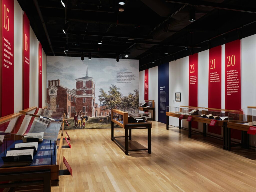 A photo of a museum gallery with a colorful enlarged reproduction of a print on one wall, and display cases of documents with explanatory wall text.