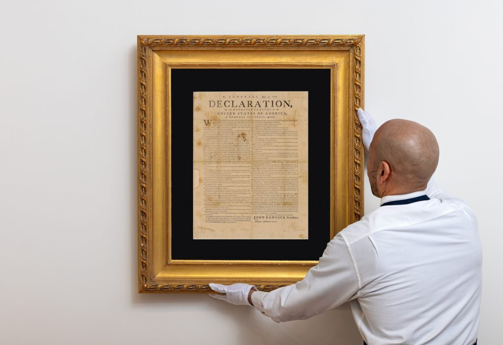 A photo of a man in white gloves carefully hanging a framed printed copy of the Declaration of Independence from July 1776. 