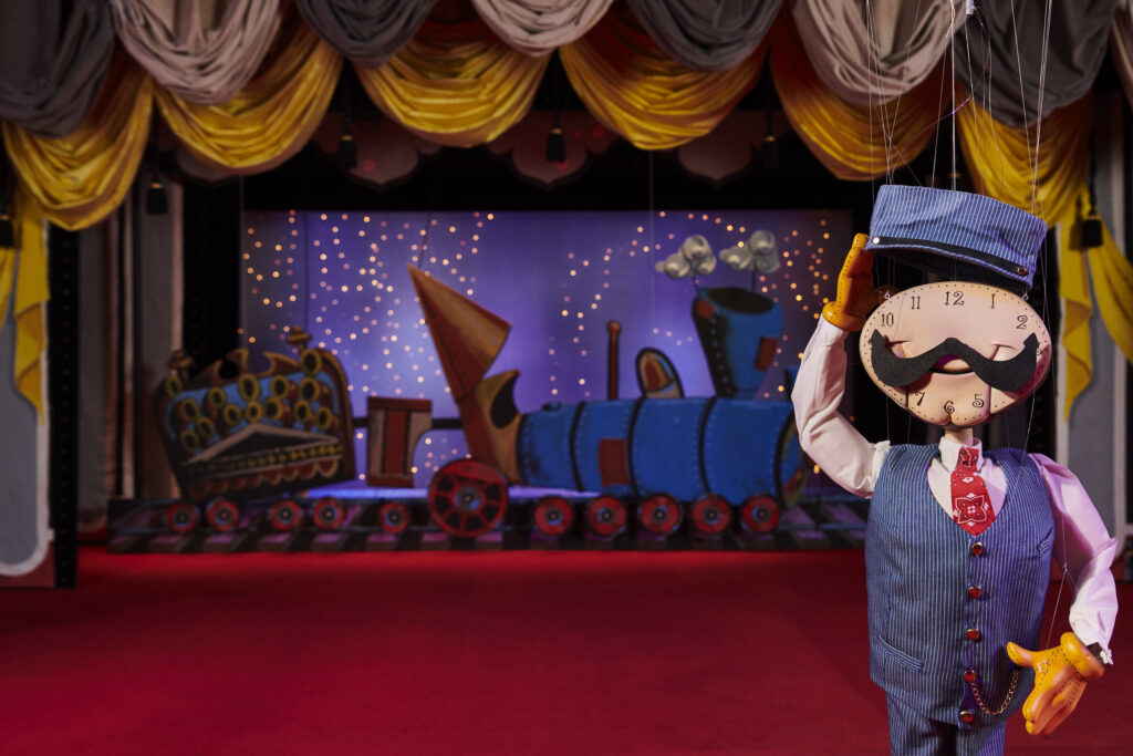 A photo of a theater stage with red curtains, with a marionette of a train conductor with a giant mustachioed clock for a face standing in front of a train set.