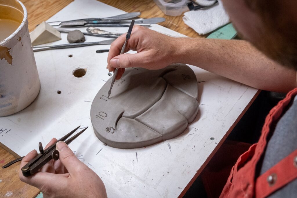 A close-up photo of the making of a clay sculpture of an anthropomorphic clock face, showing the artist's hands from above. 