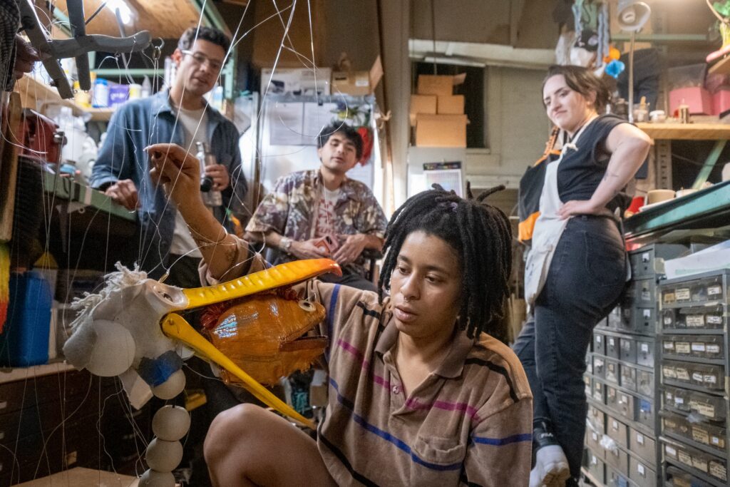 A photo of artists in a workshop looking at a marionette of a pelican with a fish in its mouth. 