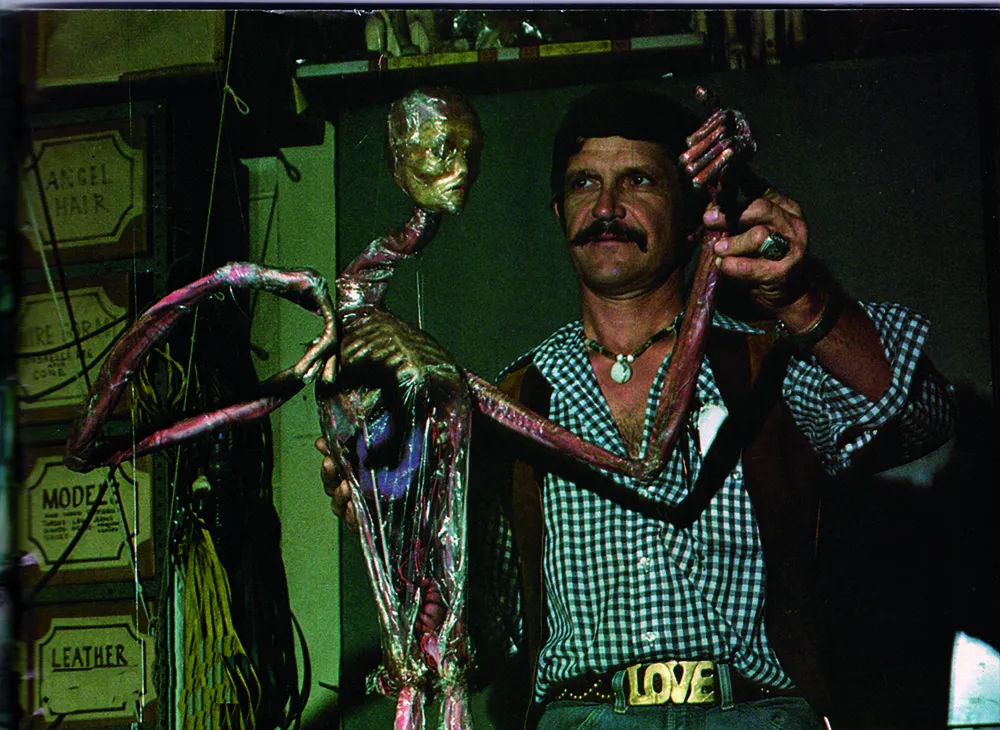 A vintage photo of a mustachioed man in a gingham shirt, a vest, and jeans with a belt buckle reading "LOVE" holds a large, creepy-looking alien puppet.