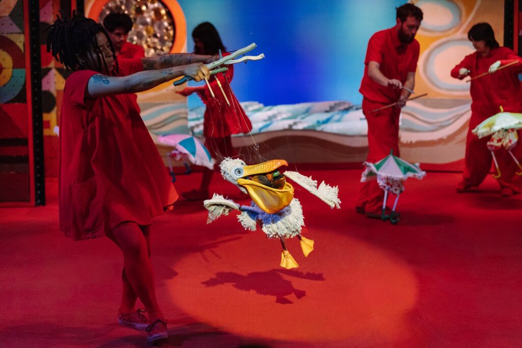 A photo of puppeteers performing on a red-carpeted stage, with a pelican marionette in the foreground. 