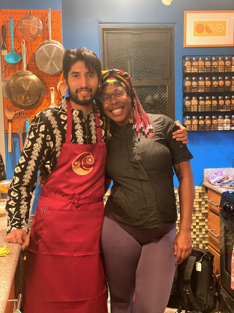 A photo of a young Latino man in a scarlet apron over a striped button-down shirt and a young black woman with pink braids in a black chef's coat standing arm-in-arm in a home kitchen with blue walls.