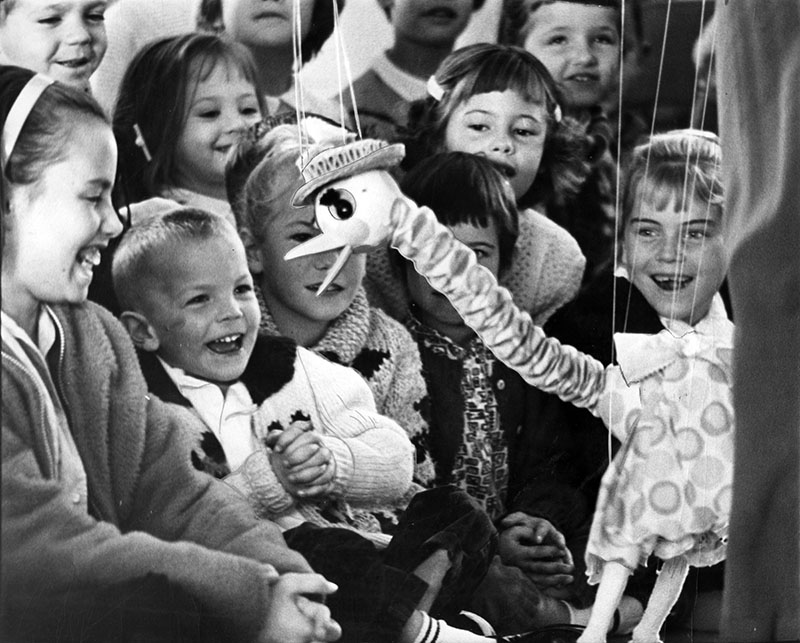 A vintage black and white photo of young children smiling delightedly at a marionette of a bird with a long neck wearing a hat. 