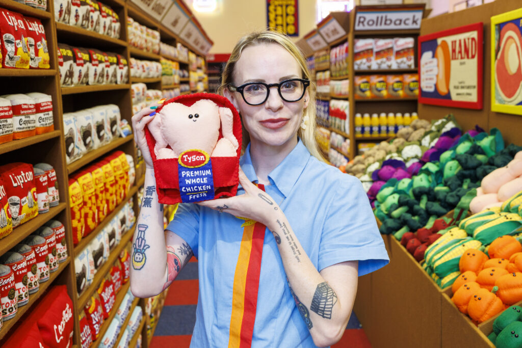 A photo of a young woman standing in an art installation of a supermarket made of felt, holding a stuffed raw chicken.