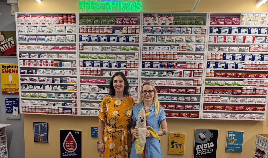 Two young women stand in front of an art display of a felted pharmacy counter.
